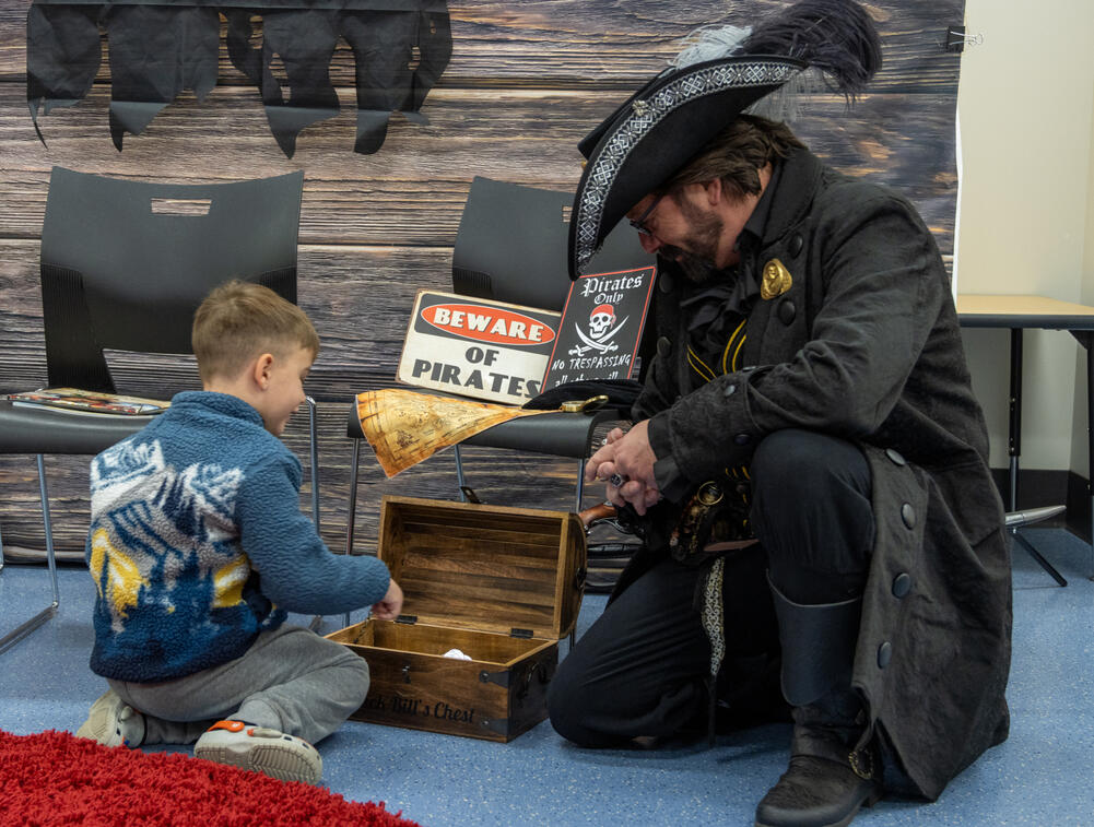 A man dressed as a pirate kneels down next to a child who is looking through a toy treasure chest